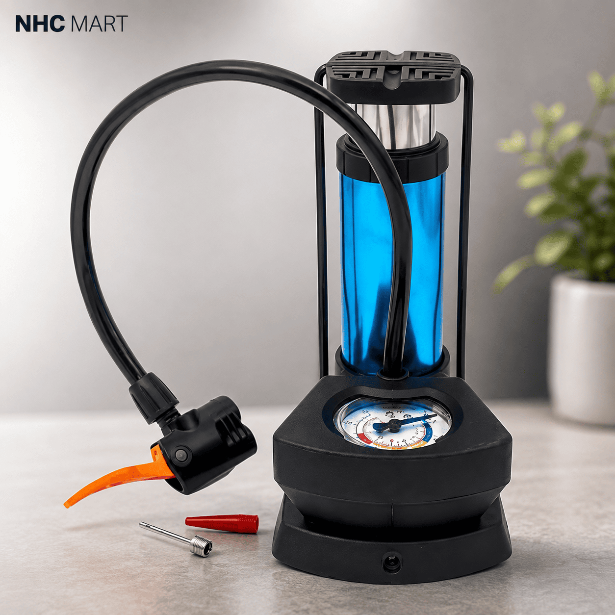 A portable bicycle pump with a black frame and hose, a translucent blue cylinder, a black base with a built-in gauge, and an orange valve connector, shown on a light gray tabletop with a small plant in the background. NHC MART logo is visible in the top left.