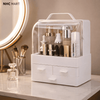 White makeup organizer with a clear plastic top cover and three drawers, shown on a marble vanity counter with cosmetics inside. A round mirror and perfume bottle are visible in the background.