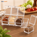Clear plastic spice container set with six square compartments, shown open on a wooden table with various spices inside. One container is detached and placed in front with its lid open.
