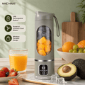 A portable electric blender in a sage green-gray color stands on a countertop with fruit and a glass of orange juice around it. The blender has a clear circular window showing blended fruit inside, a small digital display, and a carry strap.