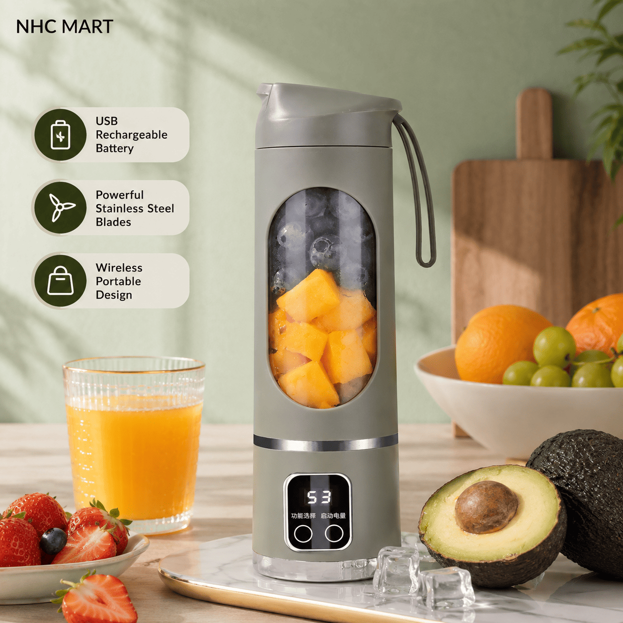 A portable electric blender in a sage green-gray color stands on a countertop with fruit and a glass of orange juice around it. The blender has a clear circular window showing blended fruit inside, a small digital display, and a carry strap.