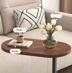 An oval wooden side table with a dark wood finish and black metal legs, styled in a living room setting on a beige sofa. The table is decorated with a clear ribbed glass flower vase containing white tulips, a brown ceramic mug on a book, and a stack of books.