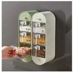 A wall-mounted spice rack with two cream-colored cabinets and six clear plastic spice jars with gold-tone handles. A hand is shown pulling out a jar of pink seasoning.